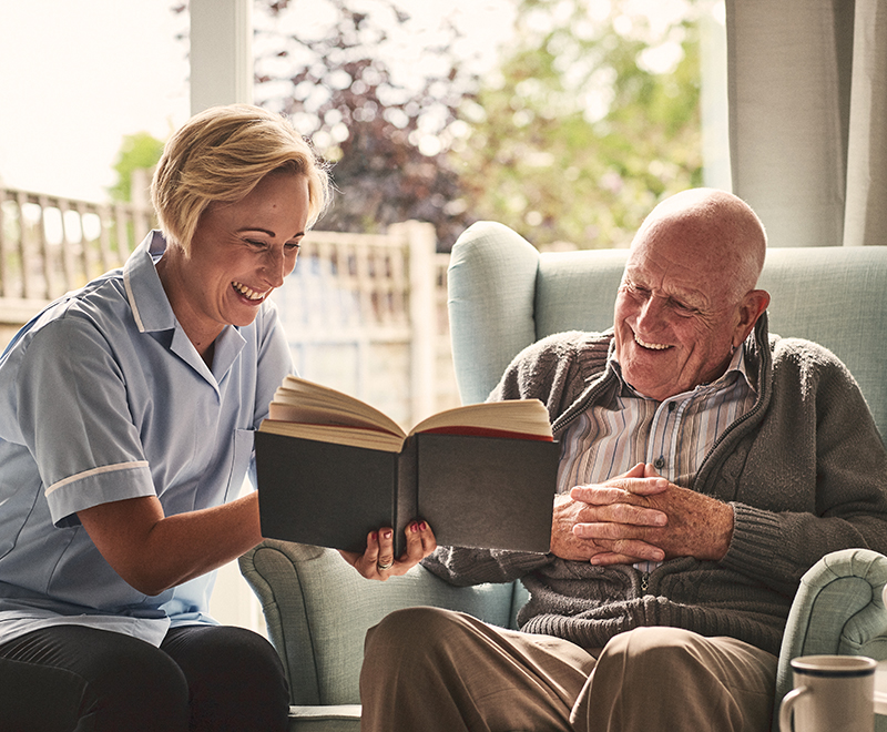 A caregiver reads a book with an elderly male patient in his home.